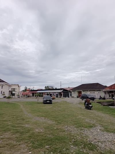 The image depicts an outdoor scene in the city of Mesjid Reuntoh, Indonesia. A mostly overcast sky covers the area. In the foreground, there is a grassy field with patches of gravel. A dirt path curves through the grass. In the middle ground, several buildings are visible. To the left, a white building with multiple windows and pillars stands. Adjacent to it is another light-colored building. In the center, a building with a reddish-brown domed roof, possibly a mosque, is prominent. Further to the right are more low-rise buildings with dark roofs.
Several vehicles are present. A black SUV is parked near the buildings. A person is riding a black motorcycle in the middle ground, moving away from the viewer. Other motorcycles are parked or being ridden by individuals in the vicinity of the buildings. People are also seen walking near the structures. A trolley loaded with goods is positioned next to the black SUV. The environment appears to be a mixed-use area with buildings, open spaces, and evidence of local transportation.