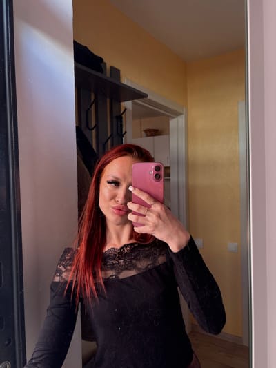 A woman with auburn hair and wearing a black lace top is taking a selfie in a mirror using a pink smartphone. The background appears to be a room interior with a yellow wall and a doorway leading to another room. A bed frame is partially visible on the left side of the mirror. The location is Simferopol, Ukraine.