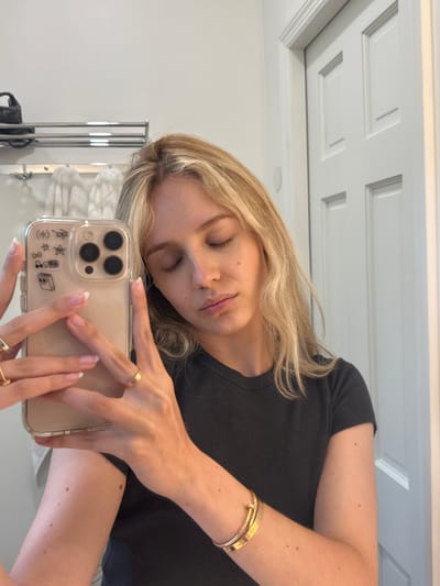 A young woman with blonde hair is taking a mirror selfie in a bathroom. She is wearing a dark grey t-shirt and several pieces of gold jewelry, including rings and a bracelet. Her eyes are closed and her lips are slightly pursed. The bathroom appears to be modern and clean, with white tiled walls and a silver towel rack visible in the background. A white door with paneling is also visible to the right of the frame. The lighting is soft and natural, suggesting it is daytime. There is no indication of the city of Miami or the United States of America in the image.