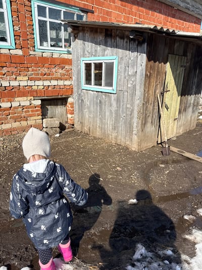 The image shows a young child standing in a muddy area near a brick and wood building. The child is wearing a patterned blue coat, a light-colored knit hat, polka dot leggings, and pink boots. The ground is a mix of mud, water puddles, and patches of snow.
The building has a section with exposed red bricks, some areas of which are deteriorating, with window frames painted light blue. Adjacent to the brick part is a wooden structure with a window and what appears to be a barred-up opening. A shovel leans against the wooden wall.
The location is presumably Кипчак, Russia, as specified. The weather appears to be cold and wet, likely early spring, judging by the melting snow and mud.