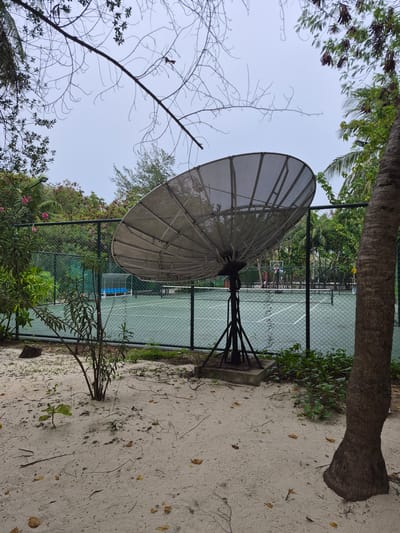 The image shows a satellite dish antenna located near a tennis court on the island of Huraa, Maldives. The antenna is a mesh type, supported by a metal frame and is positioned on a concrete base. A chain-link fence separates the antenna from the tennis court. A tree is next to the antenna, and a sandy ground covers the area. The tennis court is visible in the background with a net in the center and tennis equipment. The sky is overcast.