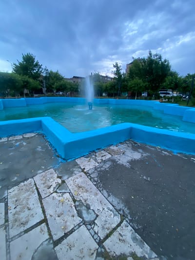 The image depicts an outdoor public space in Vagharshapat, Armenia, centered on a large, multi-tiered fountain. The fountain's concrete basin is painted a vibrant cerulean blue, and its interior holds turquoise-colored water from which a single, strong vertical jet of water ascends. The ground immediately surrounding the fountain is a mix of irregularly shaped, light-grey paving slabs, some cracked and showing signs of wear and sparse vegetation growth, and a darker, asphalt-like surface. Blue paint splatters from the fountain are visible on both paving types. In the mid-ground and background, numerous deciduous trees with dense green foliage line the area, partially obscuring multi-story buildings and parked vehicles, including a white van. The sky above is heavily overcast with layers of grey and bluish clouds. No individuals are present in the immediate foreground or mid-ground.