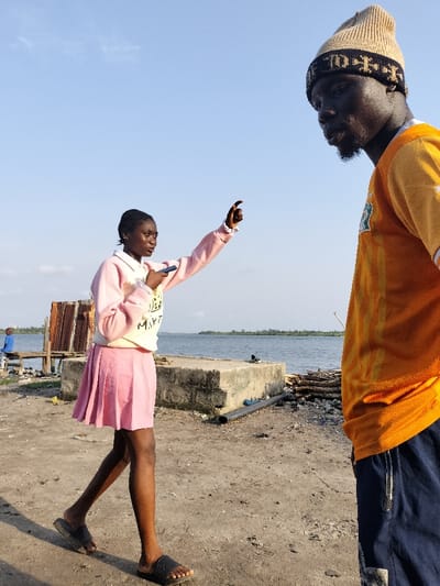 The image captures an outdoor scene on what appears to be a sandy or dirt shore beside a wide body of water, likely a river or lagoon, under a clear blue sky. The lighting suggests either late morning or late afternoon, with bright, natural light.

Two individuals are prominently featured. In the left-center of the frame, a young Black female walks towards the right side of the image, with her left foot lifted in mid-stride. She is looking towards the right and her right arm is extended upwards and forward, with her index finger pointing. In her left hand, held close to her chest, she holds a dark-colored smartphone. She wears a light pink, long-sleeved sweatshirt over a white collared shirt, and a pleated light pink skirt that falls above her knees. Black slide sandals are on her feet. On the front of her sweatshirt, there is faint light-colored text, partially visible as "MAYT???".

To the right, a Black man is partially visible, mostly his upper body, facing towards the left and seemingly looking in the direction the woman is pointing or observing her. He has a short beard and is wearing a light brown knitted beanie with a dark patterned band. He wears an orange short-sleeved t-shirt with yellow striped or patterned details, possibly a sports jersey. The lower part of his body, dressed in dark blue shorts or pants, is cut off by the right edge of the frame.

In the background, behind the woman, a rustic structure made of wood and corrugated metal is visible. A person in a blue shirt is faintly seen sitting on a wooden bench near this structure. Further back, along the edge of the water, several large, worn concrete blocks or slabs are present. To the right of these blocks, a pile of wooden logs or debris is stacked on the ground, and a black pipe lies horizontally. A pair of black shoes (possibly slides or flip-flops) rests on one of the concrete blocks near the water. The wide expanse of calm, blue-grey water stretches across the mid-ground, meeting a distant shoreline covered with faint green foliage under the clear sky.