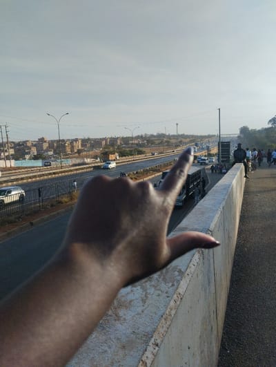 A hand with a dark nail points towards a distant highway. The highway spans the horizon, packed with vehicles and lined with streetlights. On the right side, people gather near a concrete barrier. A city skyline is visible in the background. The sky appears overcast with a slight bluish tint.