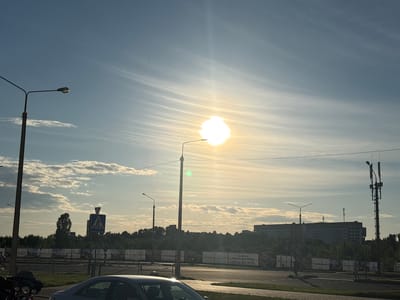 The image depicts an outdoor scene with a bright sun shining in a hazy sky, creating a lens flare effect. The sun is positioned in the upper center of the frame, casting a bright glow. Wispy clouds stretch across the sky, illuminated by the sunlight.

In the foreground, a street with a paved surface is visible. A dark-colored car is parked on the left side of the road. A pedestrian crossing sign is mounted on a pole in the middle ground, indicating a crosswalk. Further back, a line of trees forms a dense green barrier. Behind the trees, a large, multi-story building with a grid-like facade is prominent. A tall cell tower with multiple antennas stands to the right of the building.

Several streetlights are visible throughout the scene, some of them appearing to have a greenish hue due to the sunlight or lens glare. The overall setting appears to be an urban or suburban area, possibly on the outskirts of a city, given the mix of buildings, infrastructure, and greenery.

The lighting suggests it is either early morning or late afternoon. The hazy atmosphere and the sun's position imply fair weather, though not entirely clear. The presence of the car and the general urban elements suggest a typical day with no specific event unfolding.

There are several pieces of visible text in the image, predominantly on banners or signs in the background. One banner on the right reads "+375 44 525 32 70" and "Приглашаем". Another visible text includes "www.tenkin.by". These suggest commercial or informational signage. No people are clearly discernible as main subjects, though there might be very distant figures.