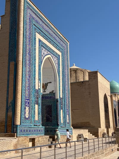 The image depicts the exterior of the Gur-e-Amir Mausoleum in Samarkand, Uzbekistan. The mausoleum is characterized by its intricate blue and turquoise tilework adorning the facade, particularly around the grand arched entrance. The brickwork of the structure is visible, contrasting with the detailed tile mosaics. In the foreground, a paved walkway is bordered by a metal railing, with several people walking and standing near benches. The sky is a clear, bright blue. Adjacent to the main mausoleum, another part of the complex, likely the mosque, is visible with a dome and brick construction. The architectural style suggests a Timurid influence, common in Samarkand.