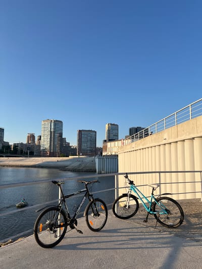 This image captures a waterfront scene in Khabarovsk, Russia, likely near the Amur River embankment. Two bicycles, a black Merida and a blue mountain bike, are parked on a paved promenade overlooking the water. In the distance, a skyline of modern, multi-story buildings dominates the view, with a clear blue sky above. A person is visible rowing a small boat on the river. The architecture suggests an urban setting, with the water and riverside infrastructure indicating a developed city. The lighting suggests it is late afternoon or early evening, with long shadows cast on the promenade.