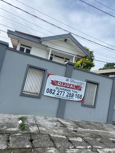 The image shows a two-story house with a gray wall. The house is located in Katapang, Indonesia, at the coordinates -6.99 latitude and 107.557 longitude. A sign with the words "DIJUAL" (for sale in Indonesian) is hanging on the wall. The sign also includes a phone number and the words "GUNAWAN TAN" and "Discovery Property" printed in red. There is a gated section of the wall, and a small amount of vegetation and trash is visible in the foreground. The house is partially visible in the background. The sky is visible at the top of the image and there are multiple power lines stretching across the sky. The photo was likely taken from the street.