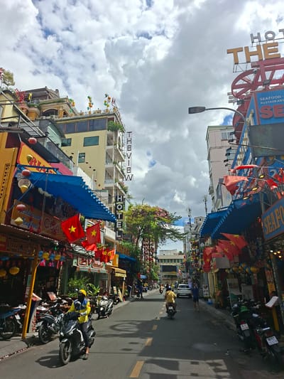 A street scene in Thủ Đức city, Vietnam, features a paved road lined with multi-story buildings adorned with flags, banners, and lanterns. The buildings house various establishments, including hotels and restaurants, with prominent signage such as "THE VIEW HOTEL" and "SEAFOOD RESTAURANT." Several motorcycles are parked along the street and in motion. One person on a motorcycle, wearing a yellow jacket and helmet, is riding towards the viewer. Another person on a motorcycle, in a yellow shirt, is riding away from the viewer. A person in a blue shirt is walking on the sidewalk. The sky above is filled with dramatic clouds.