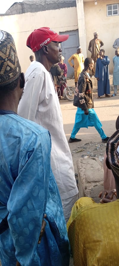 The scene depicts a daylight street gathering in Kabuga, Nigeria, with multiple individuals present. In the immediate foreground, from the left, a person wearing a blue patterned garment with circular motifs is partially visible, facing right. To their right, a man in a plain white garment and a red baseball cap, embroidered with "NNPP" in white letters on the front, stands facing right in profile. His gaze is directed forward.

In the midground, to the right of the man in white, another individual walks right to left, wearing a dark jacket adorned with light-colored patterns and bright blue loose-fitting pants, with black shoes. Behind this moving figure, a silver-colored motorcycle with a spoked wheel is parked or stationary. Further in the midground and background, several other men are visible, primarily standing. They wear traditional Nigerian attire, including various solid-colored garments such as brown, dark blue, light blue plaid, and maroon, with some wearing skullcaps.

The background consists of a light-colored building with an off-white or beige textured facade. A rectangular door is visible on the left side of this building, and a small, white-framed window is positioned higher on the right. The ground in the foreground and midground is a mix of dirt and gravel, with a concrete curb or block visible on the right. The lighting suggests a bright, possibly late morning or afternoon sun, casting soft shadows.