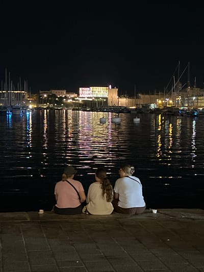 Three individuals, viewed from behind, are seated on a tiled waterfront promenade at night. The person on the left wears a pink t-shirt and a dark baseball cap. The middle person has long, brown hair and wears a light-colored shirt. The person on the right has blonde hair and wears a white t-shirt with a red strap over their shoulder. They are all facing a body of water that reflects colorful lights from buildings and boats in the background. The background shows a cityscape with illuminated buildings and numerous sailboats docked in a harbor, characteristic of the Old Port of Marseille, France. The water is calm, with ripples disturbing the reflections. Two small white cups are visible on the promenade near the individuals.