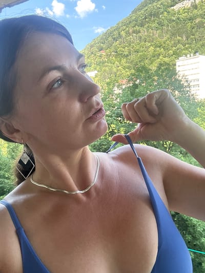 A low-angle, close-up, portrait-oriented shot shows a woman with dark hair and light skin looking up and to her left. She is wearing a blue bikini top and a silver necklace with a black, square pendant. Her right hand is reaching up to adjust one of the bikini straps. Her lips are slightly parted. The background is a blurred view of lush green trees and a light-colored building on a hillside. The sky, visible in the upper left corner, is blue with scattered white clouds. The sun appears to be shining, suggesting it is daytime. The scene conveys a casual, outdoor atmosphere.