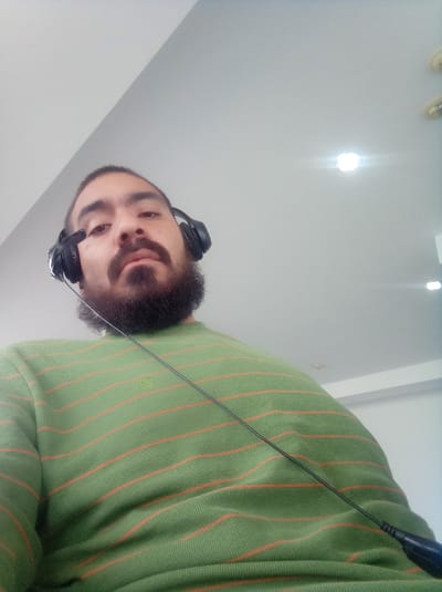 A person with a dark beard and wearing black headphones and a green and orange striped shirt is shown from a low angle. The background consists of a white ceiling with recessed lighting. The location in the city of Parroquia Guatire, Venezuela cannot be determined from the image.