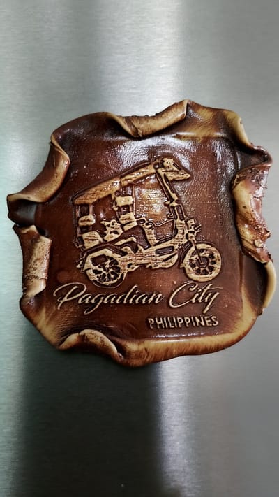 The image shows a brown, embossed souvenir magnet with an irregular, wavy border, reminiscent of tanned leather or wood. The magnet features a relief carving of a three-wheeled motor vehicle, likely a tricycle or pedicab, with a passenger compartment. Below the vehicle, in a flowing script, are the words "Pagadian City," followed by "PHILIPPINES" in block letters. The magnet is placed on a brushed metallic surface, possibly a refrigerator door, which reflects light. The background is out of focus, showing the texture of the metallic surface. There are no people, actions, or interactions depicted in the image, nor is there any connection to Smolyan, Bulgaria.
