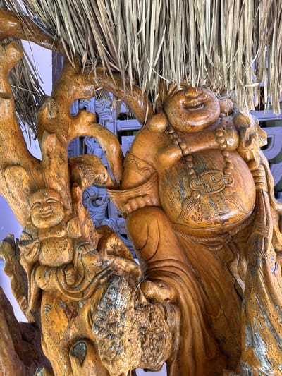 A close-up, eye-level shot captures a wooden carving of a laughing Buddha figure. The carving, made of polished wood with a warm, orange-brown hue, features intricate details such as a smiling face, a prominent belly, and draped clothing. The figure is adorned with a beaded necklace and is supported by what appears to be a carved tree trunk with a rough, textured surface. Overhead, dried straw or grass hangs down, partially obscuring the top of the carving. The background is blurred, revealing glimpses of purple and blue decorative elements, suggesting an outdoor or semi-outdoor setting, possibly in Thôn 5, Vietnam.