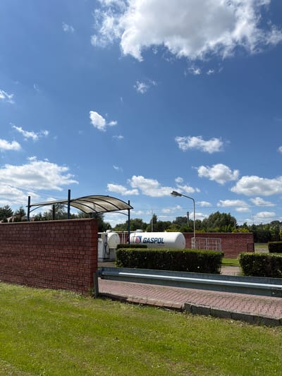 The image captures an outdoor scene on a sunny day with a blue sky and scattered white clouds. In the foreground, a green lawn is visible, bordered by a red brick wall on the left and a metal guardrail on the right. Beyond the wall and guardrail, a paved pathway is laid with red bricks.
The main subjects are two large white cylindrical tanks. One of the tanks, partially visible on the left, has a white dispensing unit attached to it. The other, larger tank is prominently displayed with the blue and red "GASPOL PROPAN" logo. These tanks are positioned behind a low, manicured hedge.
A clear plastic canopy supported by metal poles stretches over part of the area near the dispensing unit. A street lamp with a curved arm stands in the middle ground to the right. In the background, trees and foliage form a dense line, with some houses and utility poles visible interspersed. The scene appears to be a gas station or a storage facility for propane.
The location is identified as Strzyżowice, Poland. The overall impression is one of an operational industrial or service area under a bright sky.