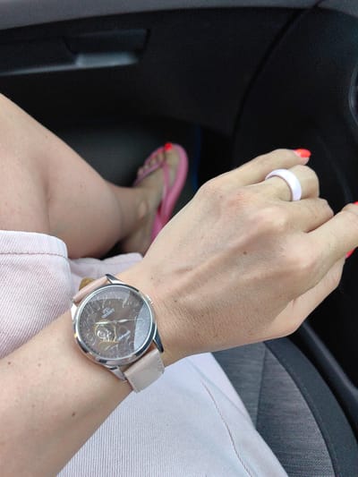 A woman's arm, wearing a silver-toned watch with a light pink band and a white ring, is visible in the foreground. Her nails are painted a bright orange color. The watch face is dark brown with silver hour markers and visible internal mechanism. The background shows a woman's legs in pink flip-flops and a light pink skirt, with a dark interior of a vehicle. The image is taken from a first-person perspective, likely inside a car, with the woman's arm resting on the vehicle's seat. The scene appears to be in the city of Дзяржынскі сельскі Савет, Belarus, though no specific landmarks are visible.