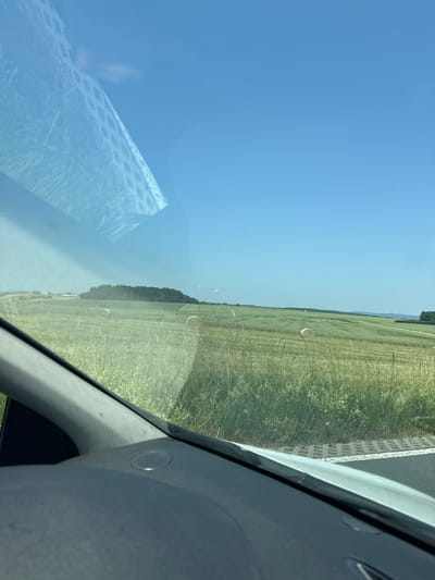 The image is taken from inside a vehicle, looking out through the windshield at a rural landscape under a clear blue sky. The foreground shows parts of the vehicle's interior, including a dark dashboard and a portion of the windshield frame. Reflections are visible on the windshield, particularly a lattice pattern.
The midground and background depict a rolling green landscape. There are fields of grass and possibly crops, with some areas appearing lighter green and others darker. In the distance, there are several round hay bales in the fields. A treed hill or small mountain is visible on the left side of the image. Further in the distance, on the right, what appear to be wind turbines are present on the horizon, along with some buildings and a road or path. The environment appears to be agricultural and open countryside, likely in Germany given the specified location. There are no people or animals directly visible in the image. The overall impression is a sunny day in a rural setting.