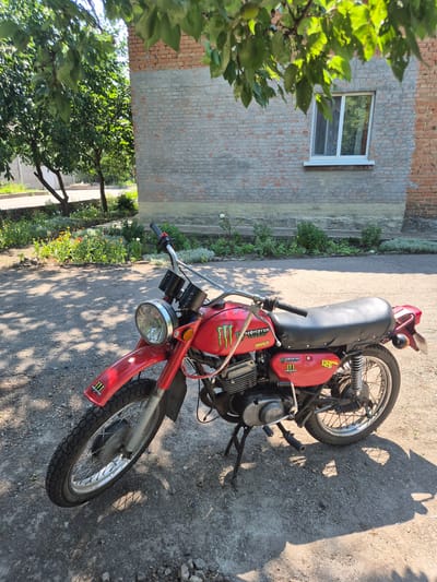 In the village of Sozonivka, Ukraine, a red vintage motorcycle with "Monster Energy" branding is parked on a gravel and asphalt surface in front of a brick building. The motorcycle is positioned at an angle, with its front wheel turned slightly to the left. It has a single round headlight, chrome handlebars, and a black seat. A kickstand supports the motorcycle. The building behind it has a light gray lower section and exposed brick above, with a white-framed window visible. Lush green trees with dappled sunlight casting shadows on the ground are present in the foreground and overhead. The surrounding area shows a flowerbed with various colorful flowers and greenery, suggesting a residential or rural setting. The sky is bright and clear, indicating a sunny day. There are no people or animals visible in the image.