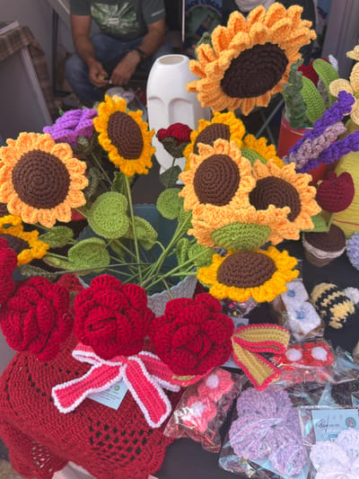A crocheted flower arrangement, featuring sunflowers and roses, is displayed on a table in a shop in Sector 17, Chandigarh. The sunflowers are made of yellow yarn, with brown centers, and the roses are made of red yarn. The arrangement is placed in a blue bucket with green crocheted leaves, and a white plastic container sits behind it. The shop is selling other crocheted items, including a small bee, lavender flowers, and a heart-shaped crocheted blanket with a white ribbon. The scene is a close-up view of the crocheted arrangement and surrounding items. The background is out of focus.