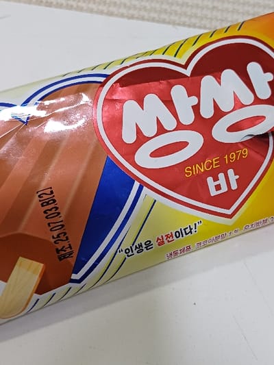 A single wrapped Ssaang Ssaang Bar (쌍쌍바) ice cream is displayed on a light-colored, flat surface. The product packaging is oriented diagonally across the frame, from bottom-left to top-right. The wrapper's design features a prominent red heart-shaped logo with the Korean text "쌍쌍" in white, and "SINCE 1979" with "바" (Bar) in yellow beneath it. The left portion of the wrapper is brown, indicating the flavor, and includes a visible wooden popsicle stick extending from the bottom-left edge. Black text on the brown section reads "제조.25.07.03.8(2)", consistent with a manufacturing or expiry date (July 3, 2025, batch 8, item 2). The right portion of the wrapper is yellow with horizontal blue and white stripes, bearing the Korean phrase "인생은 실전이다!" (Life is a real experience!) in black text, along with smaller, partially visible Korean text. The immediate background is a plain, light-colored surface, while the upper-right background shows a subtly textured, light-colored wall or fabric, slightly out of focus. The wrapper exhibits minor creasing. The image depicts this scene within 서대문구, South Korea.