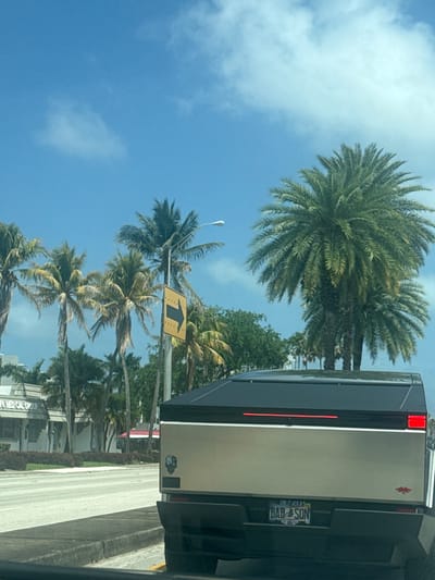 The image shows a view from a vehicle, likely a car, on a road in North Miami Beach, Florida. Palm trees line the street against a blue sky with scattered clouds. A road sign with a left-pointing arrow is visible among the trees. The rear of a gray pickup truck is prominently in the foreground. The license plate on the truck reads "BAB4SON". A small sticker of a skull is seen on the lower left part of the truck. The road appears to be paved and has a concrete curb. The building "APA MEDICAL GROUP" is seen among the trees.