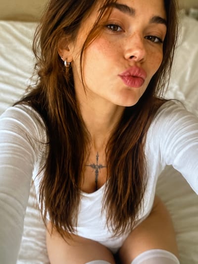The image is a close-up, selfie-style portrait of a young woman with long, dark brown hair, freckles, and dark brown eyes. She is wearing a white, long-sleeved top and has a cross tattoo on her upper chest. She is puckering her lips as if for a kiss. The background appears to be a bed with a white textured duvet. The image does not provide any information about the location in the city of Neubrandenburg, Germany.