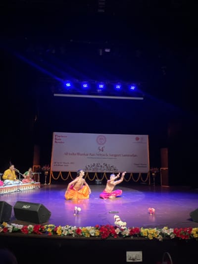 The image shows two men in orange and red traditional Indian clothing performing a dance on a stage. The stage is decorated with flowers and has a white banner that says "54th All India Bhaskar Rao Nritya & Sangeet Sammelan". The event took place at the Tagore Theatre in Chandigarh, India, located at latitude: 30.733, longitude: 76.789. There are two additional men in the background playing instruments. Above the stage is a row of blue LED lights. The stage is illuminated with blue spotlights. The background appears to be dark and empty except for the banner and a speaker on the left side of the stage.  The image is taken from the audience area, giving a slightly elevated view of the stage.