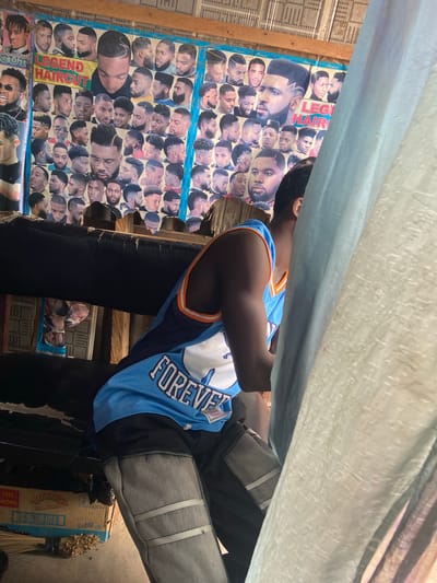 The image shows a person wearing a blue and orange basketball jersey with "FOREVER" printed on it, and gray pants. They are sitting down, with their back to the camera, in what appears to be a barbershop or a similar establishment in Kuson, Nigeria. The walls of the establishment are decorated with posters displaying various hairstyles on different men. The posters are mostly in a collage format, with smaller images of haircuts arranged around larger ones. Some of the text visible on the posters includes "LEGEND HAIRCUT" and "CUT". The setting appears to be outdoors or in a semi-enclosed space, with a light blue tarp or fabric overhead, suggesting it might be a temporary setup or an open-air business. The lighting suggests it is daytime. The overall impression is that of a bustling local business showcasing its services.