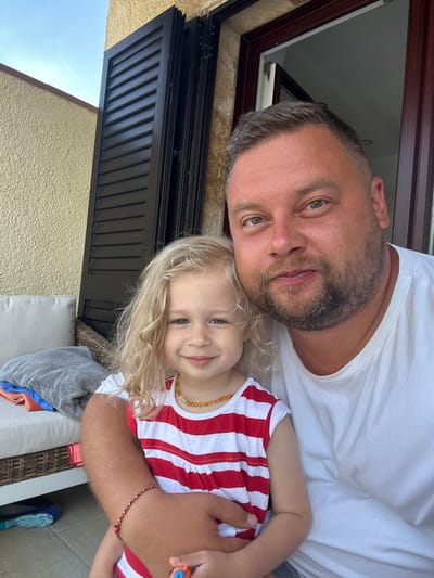 The media file shows a selfie of a man and a young child outdoors. The man has short, graying hair, a beard, and is wearing a white t-shirt. The child, with blonde hair and blue eyes, is wearing a red and white striped shirt and a beaded necklace. The child is nestled in the man's arms, and both appear to be smiling.
The background suggests an outdoor setting. There is a textured yellow wall, black louvred shutters on a door or window, and a glimpse of a blue sky. The lighting indicates it is daytime, likely sunny. The overall mood is affectionate and relaxed. A woven basket, cushions, and cleaning tools, including a blue brush, are visible on the ground, suggesting a patio or backyard area. The location context is Sant Feliu de Guíxols, Spain. No visible text is present in the media.