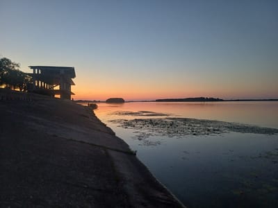 At sunset, a man-made concrete embankment slopes down to a calm body of water, likely the Dnieper River near Ukrainka, Ukraine. On the embankment, a dark, silhouetted structure with multiple vertical supports and a flat roof stands against the colorful twilight sky. The water's surface reflects the orange and blue hues of the sunset, with scattered patches of lily pads dotting the surface. In the distance, a tree-lined shore forms a dark horizontal line against the horizon. A small, dark boat is visible near the shore, and a few small figures are present on the embankment and near the water's edge. The sky transitions from a pale blue overhead to a warm orange glow near the horizon.