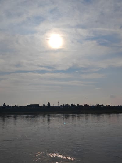 The image depicts a tranquil scene of a wide river with a village lining the opposite bank, under a sky filled with scattered clouds and a bright sun. The sun, partially obscured by clouds, is positioned high in the sky, casting a radiant glow that reflects on the water's surface. The river is calm, with gentle ripples disturbing its dark, reflective surface.

The village is silhouetted against the sky, appearing as a dark line of buildings and trees on the horizon. Among the structures, several houses of varying sizes are visible, along with what appears to be a water tower, suggesting a rural or semi-rural setting. The trees are sparse and appear to be deciduous. The overall atmosphere is peaceful and serene, evoking a sense of quietude. The village is located in Yukséevo, Russia.