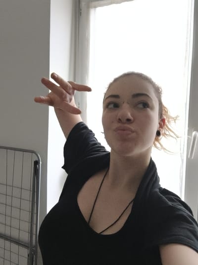 The scene is an interior shot, located in the city of La Madeleine, France. A young adult female is depicted from the mid-torso upward, positioned slightly right of center, with her head turned and eyes directed towards the upper right. Her lips are pursed in a puckered expression. She has light-toned skin, light brown or blonde hair, a bridge piercing on her nose, and a black circular ear gauge in her visible left earlobe. Her right arm is raised, with fingers extended and splayed. She wears a black scoop-neck top with visible thin straps, overlaid by a loose black top or cardigan. A thin black cord necklace rests on her chest. To the left of the subject, a chrome-plated metal garment rack or shelving unit with a grid pattern is partially visible. The background consists primarily of a brightly illuminated window with a white frame, indicating an outdoor light source beyond.