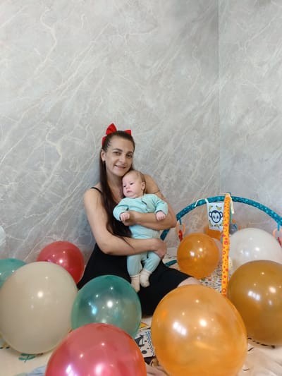 A medium shot depicts a woman holding a baby, surrounded by colorful balloons. The woman, with long dark hair adorned with a red bow, smiles at the camera. She wears a black sleeveless top. The baby, dressed in light blue, is held securely in her arms. A baby play gym with hanging toys is partially visible to the right. Several balloons in shades of red, gold, white, and teal are scattered around the floor. The background is a light grey wall. The location is inferred to be Andreyevo, Russia, based on the source information.