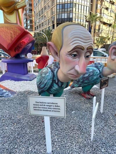 The image shows a large, colorful sculpture of two figures with human heads and bird-like bodies, situated outdoors on a bed of gray gravel. The figure in the foreground has a distinctly pained expression on its face, with wrinkled skin and downturned eyes, and appears to be composed of segmented, feathered wings. A sign in front of this sculpture reads in Spanish, "Dos buitres carroñeros tienen sed de sangre y dolor, pues los vemos muy enteros mientras siembran el terror." This translates to "Two scavenger vultures are thirsty for blood and pain, as we see them very whole while they sow terror." To the right of the main sculpture, a smaller sign depicts a skeletal figure and the word "LA MUERTE," meaning "DEATH." In the background, modern buildings and palm trees suggest an urban setting. The overall scene is likely part of a "Fallas" festival, a traditional celebration held in Valencia and other parts of the Valencian Community, including Alicante, in March, characterized by the construction and public burning of large, satirical sculptures called "ninots."