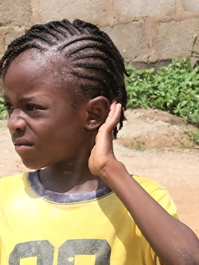 A close-up shot depicts a person with dark skin and braided hair. The individual is wearing a yellow shirt with dark blue trim and the number "6" visible on the front. The person is outside in an area with a concrete or stone surface and greenery growing nearby. The exact location in Madalla, Nigeria, cannot be determined from the image. The individual has their right hand next to their ear.