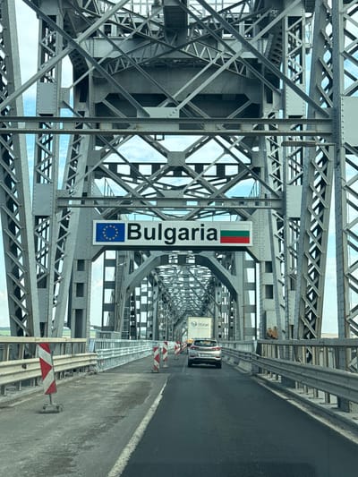 The image shows a view from a vehicle traveling on a bridge, likely over the Danube River, given the location context of Giurgiu, Romania. The bridge is a large, steel truss structure with complex metalwork visible throughout. The roadway is on the right side of the frame, with a guardrail separating it from the bridge structure. A white truck with the logo "@cargo" is visible further down the road. A sign with the text "Bulgaria" and the European Union flag is mounted on the left side of the bridge's metal structure. Red and white hazard markers are placed along the side of the road. The sky is visible through the open structure of the bridge, indicating it is daytime with some clouds. The perspective suggests the photo was taken from the driver's or passenger's seat of a car. The scene appears to be a border crossing or a point of entry into Bulgaria. There is no clear indication of people's emotions, but the overall impression is one of transit and infrastructure.