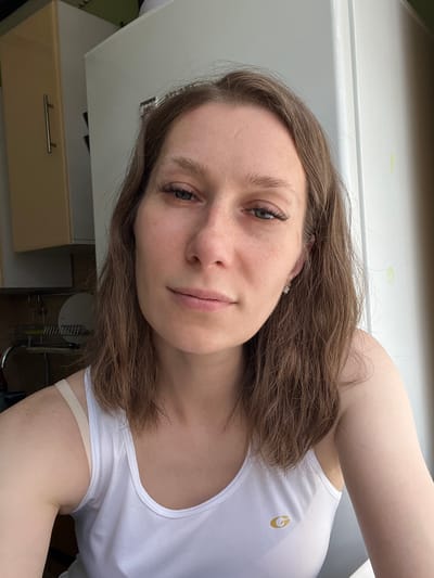 A young Caucasian woman with light brown hair is pictured in a selfie taken indoors. She is wearing a white tank top. Her hair is shoulder-length and appears slightly damp. She has applied makeup, including eyeliner and mascara, and is wearing a small stud earring in her right ear. Her expression is neutral, and she is looking directly at the camera. The background shows a kitchen area with cabinets, a sink, and a refrigerator. The lighting suggests it is daytime. This scene is likely from an apartment or house in Taldom, Russia.