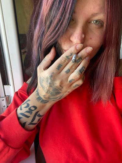 This angled, close-up portrait features a person, likely female, in Kunje, Montenegro, engaged in smoking a lit cigarette or joint. The subject has long, straight hair dyed in distinctive shades of purple, pink, and red, framing one side of their face. They are wearing a bright red long-sleeved top or hoodie, with the ribbed cuff visible on the lower right.
The person's left hand is prominent, heavily adorned with dark tattoos featuring a skull design on the back of the hand and various script-like symbols on the fingers and forearm. A ring is worn on the index finger. The fingernails are long and natural, with some slight reddish marks near the cuticles. The cigarette is held between the index and middle fingers, with a wisp of smoke visible near the nose.
Part of the person's face is visible, showing one green or hazel eye looking towards the left, alongside a portion of their nose and cheek. The expression appears neutral or contemplative. In the bottom left of the image, a white window frame is partially visible, suggesting the scene is either indoors or taken while looking out a window. No discernible text is clearly readable within the tattoos or elsewhere in the image. The scene captures a private, reflective moment.