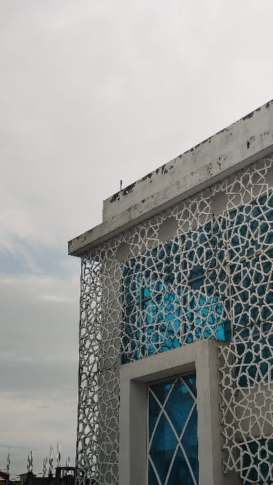 The image displays a partial exterior view of a building in Savar, Bangladesh, under an overcast sky. The upper right portion of a multi-story concrete structure is visible, featuring a flat, weathered roofline with dark stains and a slender dark rod protruding vertically. The building's facade is predominantly covered by a light-colored, ornate screen composed of interconnected geometric patterns, resembling an Islamic or Moghul jali design. This screen is positioned in front of blue-tinted glass windows, which reflect a bright, undefined light source. Towards the bottom center, a large rectangular opening, possibly a window or doorway, is framed by a solid gray concrete surround. This opening also contains blue glass, featuring a distinctive white diagonal cross pattern. The overall scene shows no discernible human activity or other prominent subjects beyond the architectural elements and the sky.