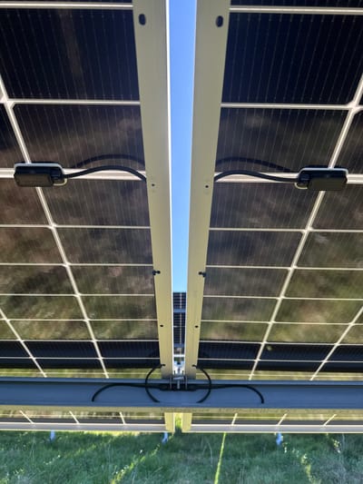 The image depicts a low-angle view of the underside of a ground-mounted solar panel array in Eastleigh, United Kingdom, under clear blue sky conditions. Multiple dark, rectangular photovoltaic panels are visible, supported by a light-colored metal framework comprising vertical posts and horizontal rails, secured with visible bolts and small black clips. Reflections of the sky and green grass below are observable on the panels' undersides. Two black, rectangular electrical components, likely power optimizers or junction boxes, are individually mounted to the underside of separate panels, positioned on the left and right, each connected by a short, curved black cable. Further black electrical wiring is routed and secured along the horizontal support beams. A distinct vertical strip of clear blue sky is visible between two central support beams, providing a view of additional, similar solar panel structures in the background distance. The scene is an outdoor installation situated over a grassy field. No personnel or other subjects are visible.