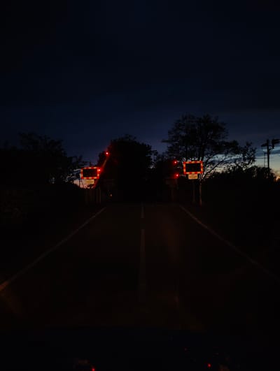 The image shows a road at night in East Suffolk, United Kingdom. Two railroad crossing signs with flashing red lights are visible on either side of the road, indicating a railroad crossing ahead. The signs read "KEEP CROSSING CLEAR." The road is paved with a white dividing line. The surrounding area appears to be wooded with trees silhouetted against the dark sky. There is also what looks like a utility pole with signals on the right side of the image. The scene is dark with the primary illumination coming from the railroad crossing signals.