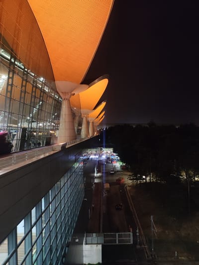 The image depicts the Kuala Lumpur International Airport (KLIA) in Sepang, Malaysia, at night. The foreground is dominated by the airport's architecture, showcasing the exterior glass walls and the iconic roof design with orange-lit, petal-like structures supported by conical pillars. Below, there's a road with vehicles and what appears to be a construction or maintenance area. Beyond the road, a dark expanse of trees is visible. The night sky is a deep black.