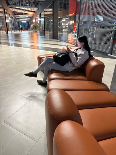 In Murino, Russia, a young woman with dark hair, wearing a light-colored blazer, grey leggings, and dark sneakers, sits on a brown leather couch in a modern indoor space. She is looking at her phone, which she holds in her hands, and has white earbuds in her ears. A black tote bag with white lettering rests beside her on the couch. The background reveals a well-lit indoor area with a polished floor, glass storefronts, and metallic ceiling structures, suggesting a shopping center or mall. The lighting creates a warm glow on the floor and highlights the architectural features of the space.