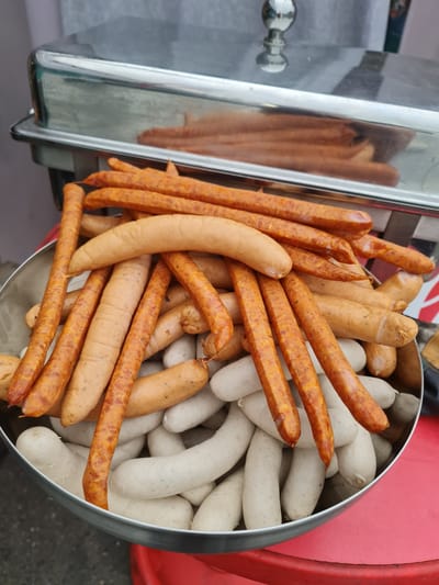 A metal serving tray containing a large pile of assorted sausages is displayed on a red surface. The sausages are of various lengths, thicknesses, and colors, with some being light beige and others a reddish-brown. The serving tray is partially covered by a metal lid with a rounded handle. The background is blurred but appears to be outdoors, possibly at a market or food stall. The context of Garbsen, Germany is not visible in the image.
