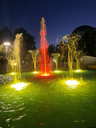 The image shows a nighttime view of a illuminated fountain in the city of Popovo, Bulgaria. The fountain features multiple water jets, some of which are lit with red and yellow lights. The water in the basin is a vibrant green color, and some koi fish are visible swimming within it. In the background, there are trees and a building with a curved roof. The sky is a deep blue, indicating it is nighttime. A lamppost is visible on the left side of the image, illuminating the trees and some people standing near the fountain.