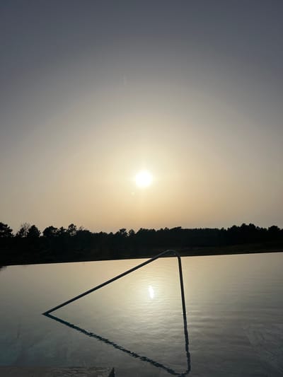 The image displays an outdoor scene at sunrise or sunset, consistent with a location in Aljezur, Portugal. The foreground is dominated by a calm body of water, likely an infinity pool, occupying the lower two-thirds of the frame. A silver-colored metal handrail extends into the water from the lower-left, curving upwards and then diagonally downwards towards the lower-right, with its reflection clearly visible on the water's surface. The water reflects the bright, circular sun positioned centrally in the sky, just above the distant treeline. This treeline, silhouetted against the sky, consists of dense, dark vegetation extending horizontally across the midground. The sky exhibits a gradient, transitioning from a bright yellow-orange hue near the horizon to a desaturated blue-grey in the upper portion of the frame. No individuals or active subjects are present in the scene.