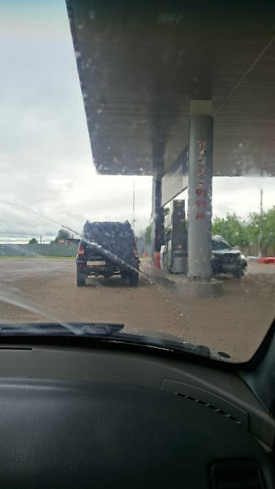 The image shows a view from inside a vehicle looking out at a gas station. The perspective is skewed, with the scene appearing sideways. A gas pump canopy extends from the left side of the frame, supported by a pole with various signs. These signs include "no smoking" symbols and fuel grades like "95" and "92". Water is visible on the windshield, suggesting rain or water being sprayed, creating a blurry, streaky effect.
Several vehicles are present at the gas station. In the middle ground, a dark-colored SUV or truck is parked, its rear visible. Further back, another vehicle, possibly a van or another truck, is also at the pumps. The ground appears to be asphalt or gravel, wet from the rain. Trees and a fence are visible in the background, suggesting an outdoor setting. The sky is overcast, indicating a cloudy day.
The time of day is not clearly discernible but the lighting suggests it is daytime. The overall mood is somewhat obscured by the water on the windshield and the unusual orientation of the image, but the presence of a gas station points to a typical scene of refueling. The location cues, such as the fuel grades and the general appearance, are consistent with a gas station in Russia. No people are clearly visible. A license plate is visible on the rear of the SUV/truck, but the text is too blurry to read.