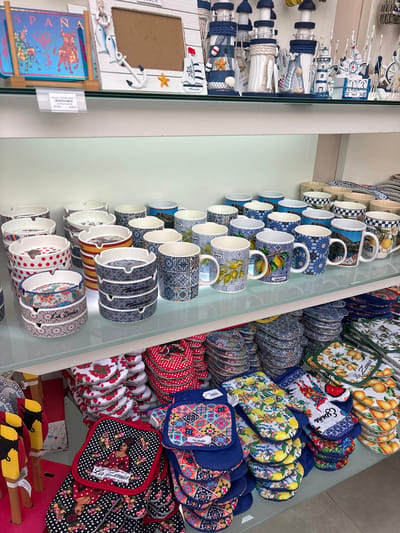 The image displays a retail display of souvenirs and kitchenware items on multiple shelves. The top shelf features nautical-themed decorative items, including lighthouse figurines and miniature sailboats, along with a framed photo holder. A price tag indicates "ESPAÑA" and "P.V.P. 4,95 €". The middle shelf showcases a variety of ceramic mugs with diverse designs, some featuring scenic landscapes, citrus fruits, and geometric patterns. Below this, several stacks of ashtrays are arranged, some with polka dot patterns and others with mosaic-like designs. The bottom shelf is filled with oven mitts and potholders, many of which are patterned with floral motifs, lemons, and Spanish-themed imagery, including one with a flamenco dancer. The items suggest a souvenir shop, likely located in a tourist area of Orihuela, Spain, given the "ESPAÑA" label and the common souvenir themes. There are no people visible in the image, and the environment appears to be an indoor retail setting.