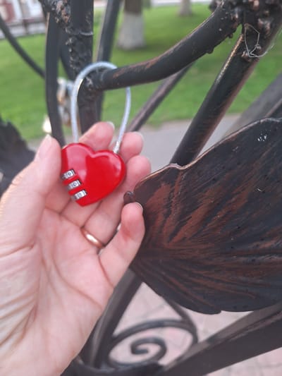 An extreme close-up shot shows a person's left hand holding a red heart-shaped combination lock. The lock has three numbered dials which are partially visible. A thin metal cable loops through the lock and attaches to a dark-colored metal fence. The fence appears to be made of wrought iron, with some decorative elements that resemble leaves. The background is blurred, but shows green grass and some out-of-focus structures, suggesting an outdoor urban environment. The image is taken from a slightly low angle, looking up towards the fence. The light suggests it is daytime, possibly late afternoon due to the warm tones on the fence.